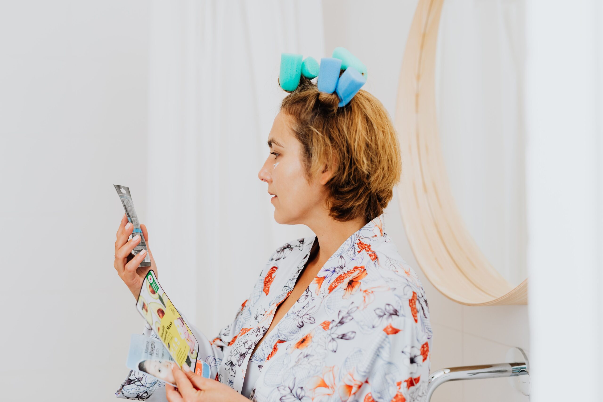 Woman in a floral robe with hair rollers reading a skincare box label