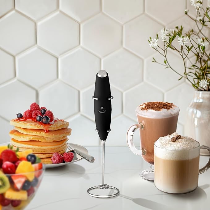 Black handheld milk frother on a small stand next to a stack of pancakes and two coffee drinks